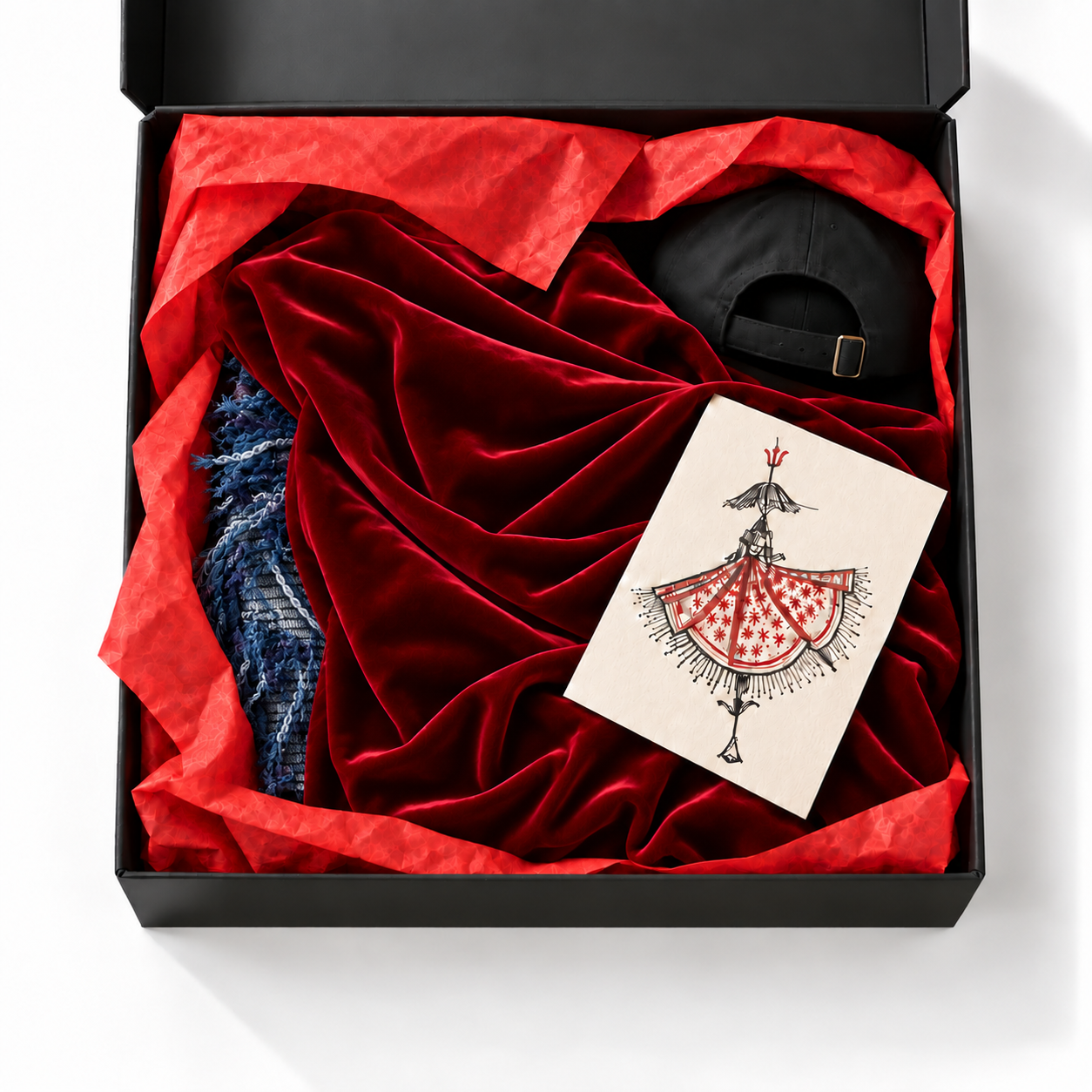 Overhead view of a black gift box with red tissue, smooth red velvet, the denim-blue Goddess Summer Top with its distinctive fringe, the back of a black cap and a sketch print. Illustrative contents.